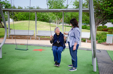 Elderly woman enjoys swinging on the swing set during a daily walk with a female caregiver.