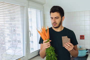 A young brunette man looks at a carrot with disdain and disgust. A person does not lead a healthy...
