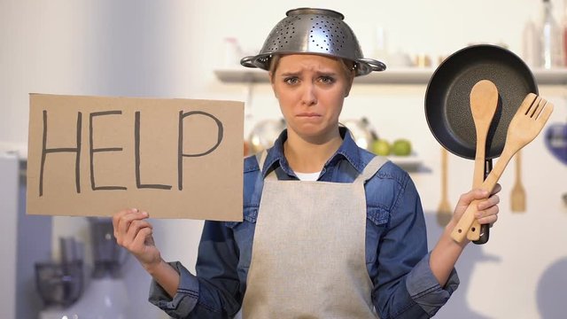 Inexperienced lady with colander on head holding kitchen utilities and help sign