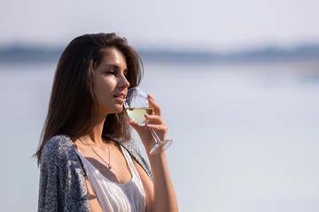 Young beautiful girl in white dress drinks wine from a glass. She is on the shore of the lake and enjoy a picnic and outdoor recreation.