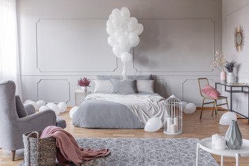 Bunch of white balloons above king size bed with grey pillows and duvet in elegant bedroom with golden chair with pink pillow and comfortable armchair