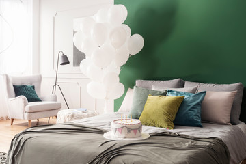 Birthday cake on double bed with grey duvet and colorful pillows in bright interior with bunch of white balloons and copy space on green wall