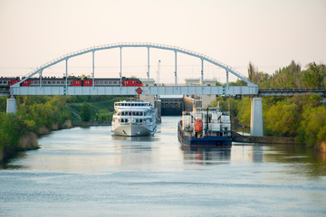 Volga-Don Canal. Volgograd. Cruise liner with tourists passes by the cargo ship and under the railway bridge on which there is an electric train.