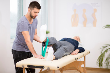 Obraz premium Grey elderly man lies on a physiotherapy table, and young doctor helps him during exercises