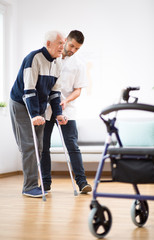 Elderly man walking on crutches and a helpful male nurse supporting him