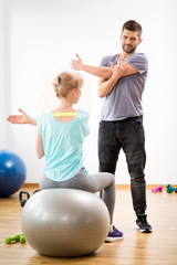 Middle age blond woman exercising on gymnastic ball during session with physiotherapist