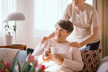 Smiling senior woman drinking tea with her caregiver standing behind her