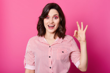 Image of attractive stylish brunette girl in striped shirt looking at camera with opened mouth and showing ok gesture, demonstrating positive emotions, being in good mood. Human facial expressions.