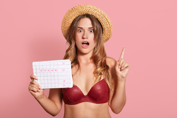 Indoor shot of shocked caucasian young female has surprised facial expression, points up with index finger, wearing in red swimsuit and straw hat, holds period calendar, forget date of periods.