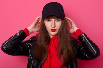 Indoor shot of young teenager wearing stylish red hoodie, leather jacket and black cap, keeps hands on head, looking directly at camera. Model posing in studio against pink wall. Teens concept.