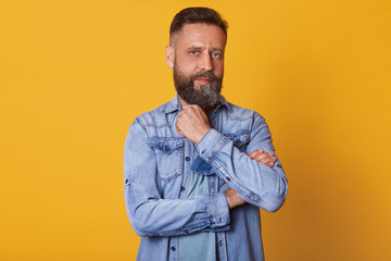 Fototapeta premium Strong thoughtful blue eyed man looking directly at camera, posing isolated over bright yellow background in studio, touching his beard with fingers, having powerful look, wearing in casual manner.