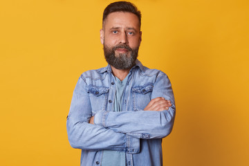 Close up portrait of bearded middle aged confident man with folded arms, standing straight, having serious facial expression, wearing casual jeans jacket, looks tired and determined. Emotions concept.