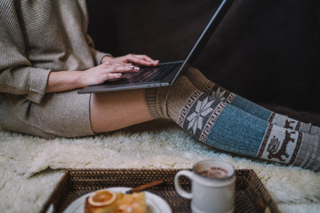 The girl sitting on a wooden floor. Laptop and coffee cup. Feet in woollen socks. The woman relaxes in cozy home. Winter and Christmas holidays concept. Selective focus. Toned image.
