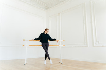 Classic ballet dancer has stretching workout, stands near ballet barres, wears black sweatshirt and leggings, does exercises in studio, focused down. Certified female instructor of choreography