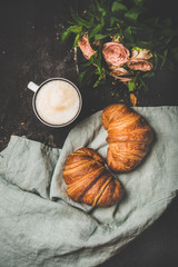 Coffee break. Flat-lay of cup of hot cappuccino with fresh croissants on grey linen towel and bouquet of pink flowers over black shabby background, top view. Spring mood