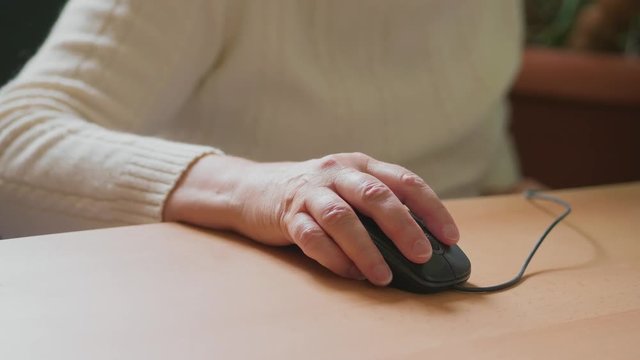 Senior woman sitting at computer browsing internet, focus moving from hand clicking on mouse to face. Grandmother looking to screen at workplace, left-clicking. Elderly lady working on laptop indoor