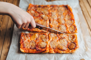 Female hand cut homemade square pizza with smoked sausage and cheese. White papyrus, wooden...