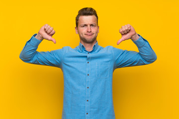 Blonde man over isolated yellow wall showing thumb down