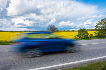 Blaues Auto fährt schnell an gelbem Rapsfeld vorüber, Schleswig-Holstein