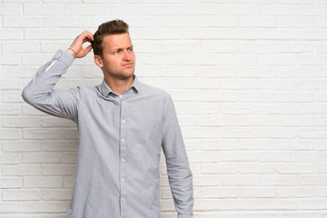 Blonde man over white brick wall having doubts while scratching head