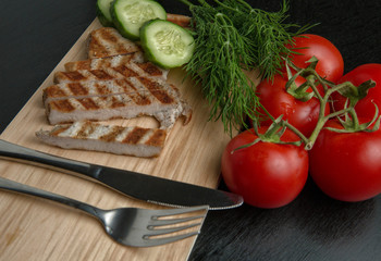 Greens with tomatoes and grilled Turkey steak on a chopping Board. Cutlery on a cutting Board. Top view