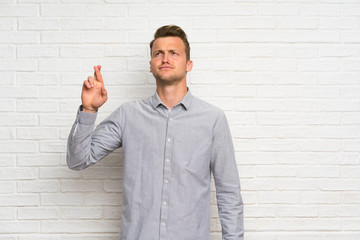 Blonde man over white brick wall with fingers crossing and wishing the best