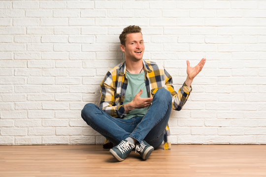 Blonde man sitting on the floor extending hands to the side for inviting to come