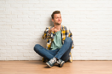 Blonde man sitting on the floor points finger at you with a confident expression