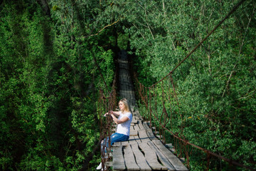 Fototapeta premium Beautiful Girl i sitting on old wooden bridge. Travel and Vacation in the park.Young blonde woman relaxing of green trees in the park. girl enjoys outdoor recreation in the spring.
