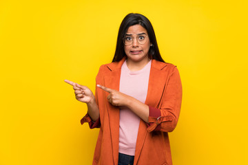 Young Colombian girl over yellow wall frightened and pointing to the side