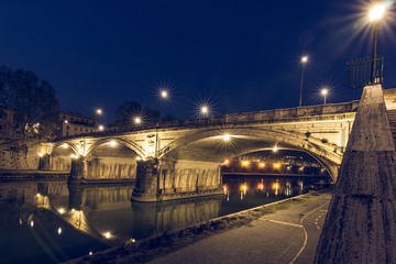 Fototapeta na wymiar Ponte Sisto is a stone road bridge in the historic center of Rome over the Tiber River at night with lighting. Path on the riverbank with lateral view at the blue hour