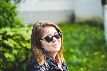 Young woman with sunglasses wearing a leather jacket smiling and feeling good in the park – Girl looking surprised in nature on a bright spring day