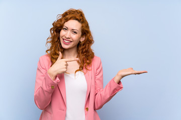Redhead woman in suit over isolated blue wall holding copyspace imaginary on the palm to insert an...