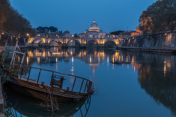 River Tiber in Rome and St Peters Cathedral at night. In the foreground a sunken ship at the pier. Buildings are illuminated. Shore with buildings and reflection of the illuminated buildings