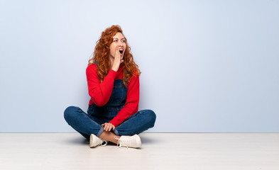 Redhead woman with overalls sitting on the floor shouting with mouth wide open to the lateral