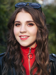 Fototapeta premium Closeup portrait of young brown-haired curly woman with trendy creative red makeup posing outdoors in bright sunlight. Pretty girl in red dress with hairstyle and makeup. 