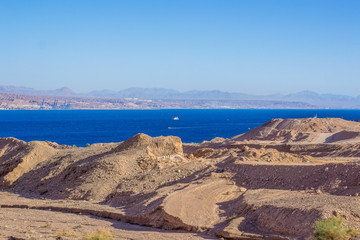 outdoor scenic landscape place from above with view on a dry sand wasteland foreground and Red sea Gulf of Aqaba with cruise ship on water urface and mountains background