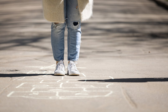 Young Woman Plays Hopscotch On Asphalt. A Girl Plays Hopscotch On A Playground In A Park Outside On A Sunny Day. Outdoor Activities For Adult And Children.