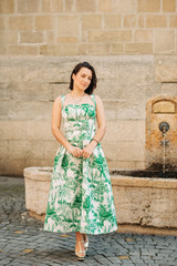 Outdoor fashion portrait of beautiful woman with dark hair, wearing long vintage styled green dress, posing on the city street