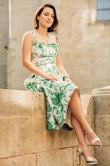 Outdoor fashion portrait of beautiful woman with dark hair, wearing long vintage styled green dress, posing on the city street, sitting on the wall