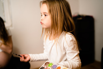 Little blondie girl holding water color palette and paint brush playing silly in front of her pregnant mother at home