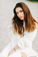 Outdoor portrait of pretty young girl wearing white pullover and jeans, sitting on the chair