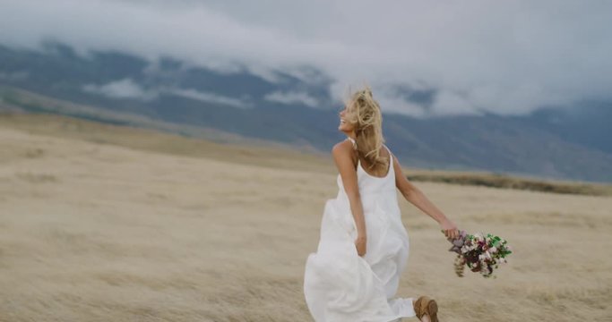 Happy laughing woman running free through a golden field in a flowing white dress holding a flower bouquet in slow motion, happy bohemian woman