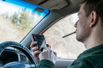 A young man inside the car with a cigarette in his mouth presses the screen of the smartphone.