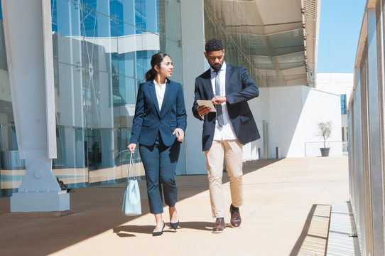 Business People Talking And Walking Along Street. Business Man And Woman Wearing Formal Clothes And Carrying Gadgets With Building In Background. Business People Concept. Front View.