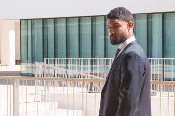 Fototapeta premium Positive ambitious business leader on his way to office. Side of young black man in formal jacket and tie walking near office building and turning face to camera. Business leader outdoors concept