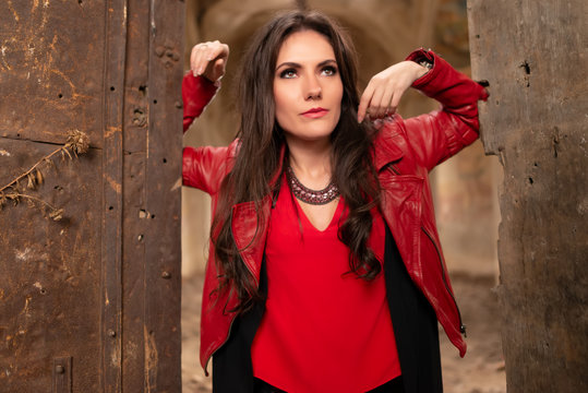 Fashion concept portrait of a gorgeous young woman in red leather jacket and black skirt outfit, sitting in a door of an old abandoned building and looking away. Body shot, retouched, vibrant colors