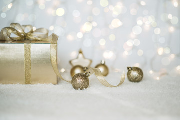 Fototapeta na wymiar Christmas background with a gold decorations and gift box in snow on the blurred, sparkling background. Toned image with copy space. Selective focus.