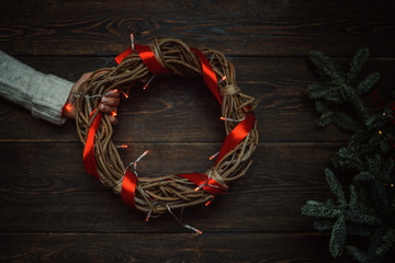 Christmas holiday background. Celebration theme. Celebration concept, free space for text. Female hand with christmas wreath with fir, pine cones, red berry and ivy on dark rustic planks background. © eskstock