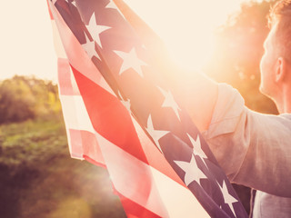 Handsome man waving an American flag against a background of trees and blue sky. View from the back, close-up. National holiday concept